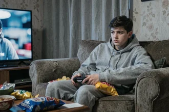 Adolescent sédentaire affalé sur un canapé avec une manette de jeu vidéo et des snacks, illustrant les risques d'obésité liés à l'inactivité physique