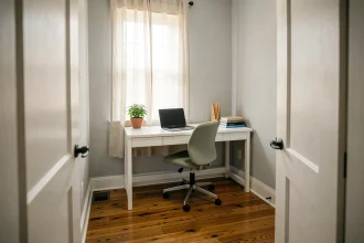 Coin bureau compact installé dans un coin de petite chambre avec bureau blanc étroit, chaise ergonomique verte et plante verte sur parquet en bois